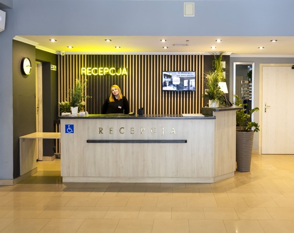 Jasna recepcja z nowoczesną drewnianą ladą, neonowym napisem i zielonymi roślinami – przytulny hotel Warszawa z profesjonalną obsługą blisko stacji metra Dworzec Wileński.
