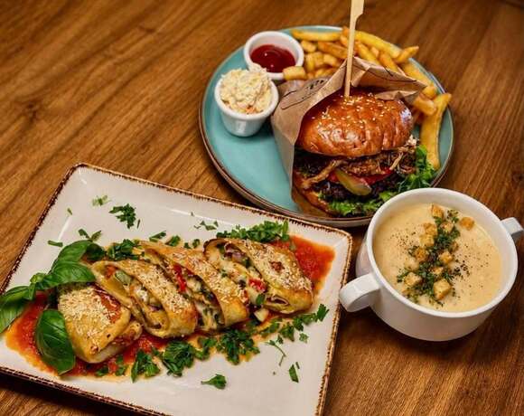 Soczysty burger gourmet z chrupiącymi frytkami i tradycyjnym colesławem, aksamitna kremowa zupa z grzankami oraz delikatne, pokrojone nadziewane naleśniki na drewnianym stole – wyjątkowe i sycące kompozycje smaków w restauracji PragaTu w Warszawie blisko stacji metra Dworzec Wileński.
