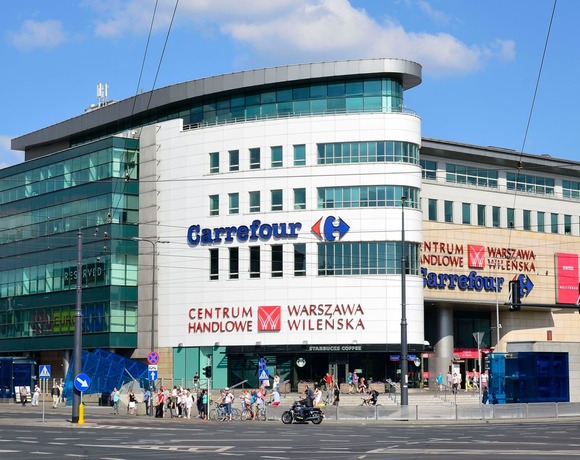 Сучасний торговий центр з вигнутою скляною фасадою та логотипом Carrefour, вуличні магазини, трамвайні дроти й перехожі