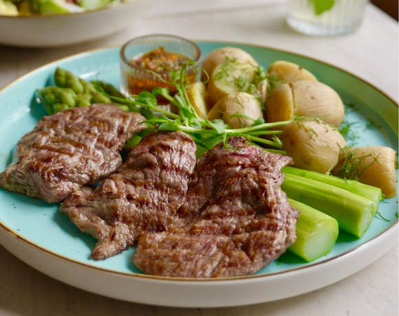 Grilled steak with new potatoes, asparagus and herb sauce on a turquoise plate, salad and mint water blurred behind