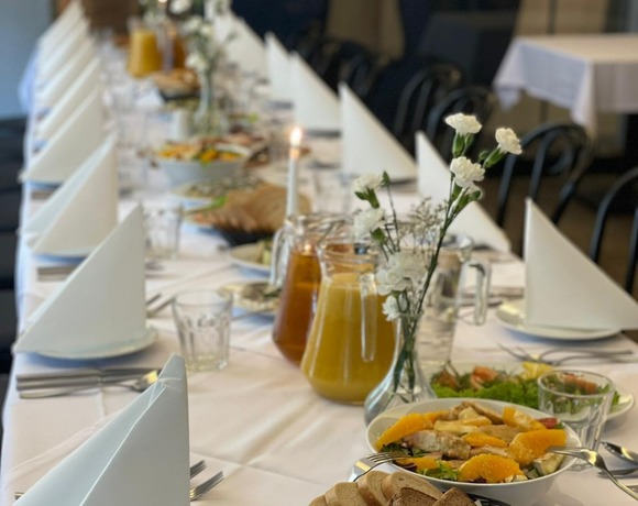 Lange Banketttafel mit weißen Servietten, Gläsern, Saftkannen, Salaten, Brotkörben, Aufschnitt und kleinen weißen Blumen