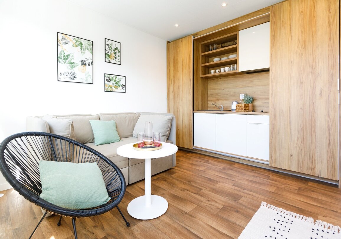 Przytulny salon z beżową sofą, zielonymi poduszkami, czarnym wiklinowym fotelem, białym stolikiem i aneksem kuchennym z drewnianymi frontami.