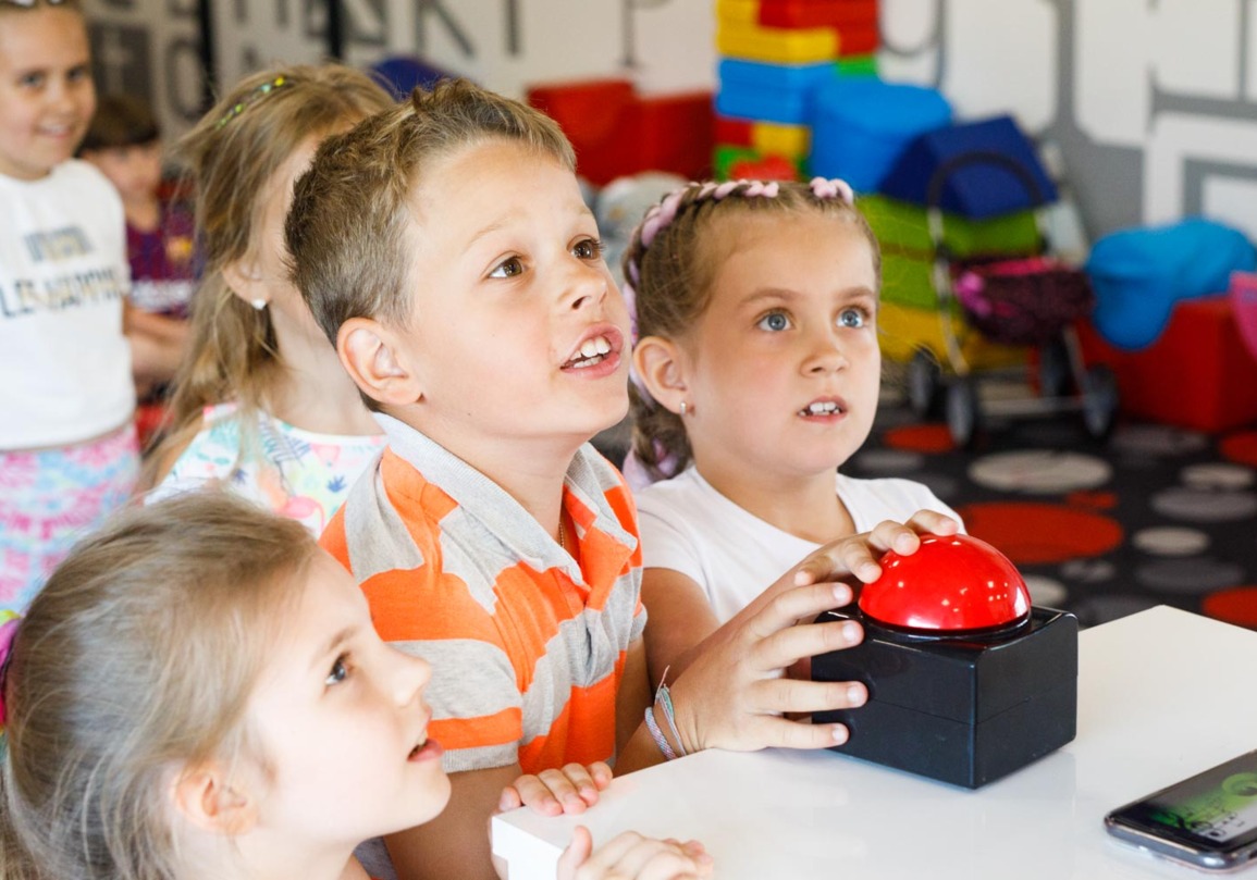 Grupa dzieci bawiących się w kolorowej sali zabaw, naciskających duży czerwony przycisk na stole.