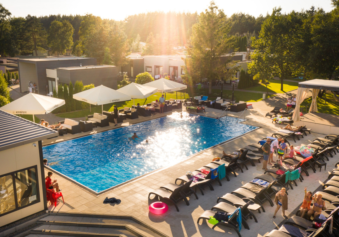 Basen z leżakami i parasolami na terenie nowoczesnego ośrodka wypoczynkowego otoczonego zielonym lasem, goście relaksują się i pływają.