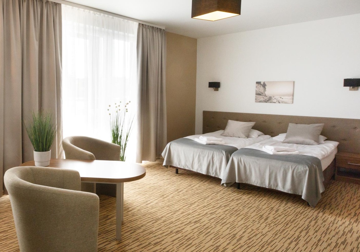 Jasny pokój hotelowy z dwoma łóżkami, beżowymi meblami i dużym oknem z zasłonami oraz roślinami