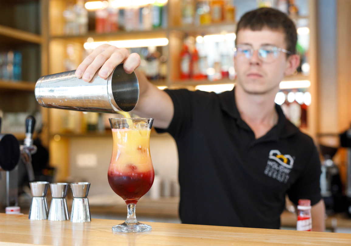 Barman w czarnej koszulce przygotowuje kolorowy koktajl w barze z oświetlonymi półkami i metalowymi miarkami