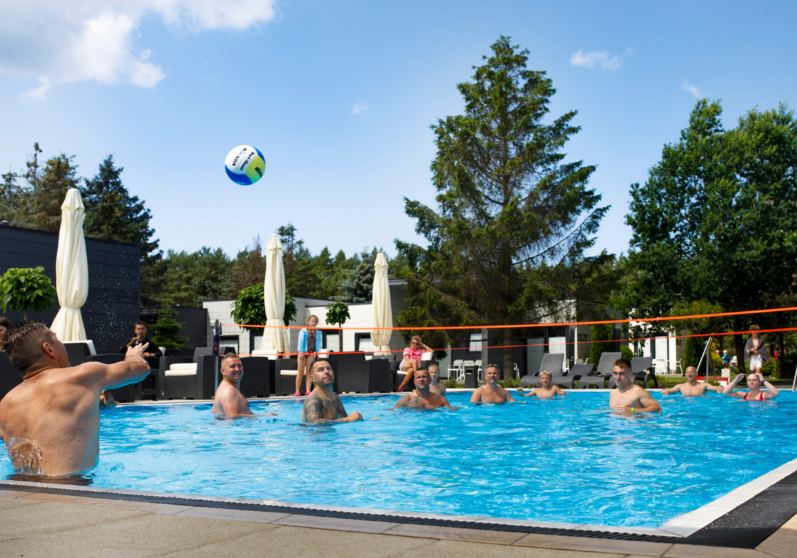 Goście grają w siatkówkę wodną w odkrytym basenie otoczonym zielenią i leżakami w ośrodku wypoczynkowym.