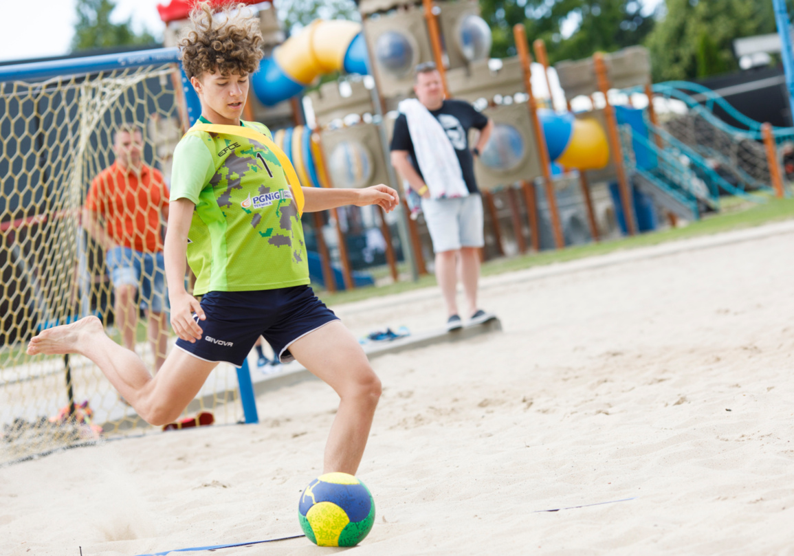 Młody chłopiec kopiący piłkę na piaszczystym boisku z placem zabaw w tle i dorosłymi obserwującymi