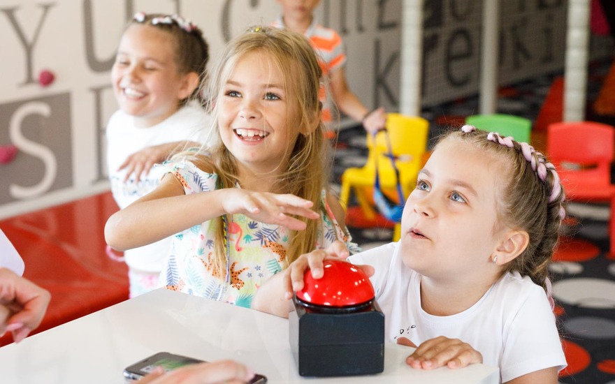 Children enjoying play area in hotel, pressing a big red button, surrounded by colorful chairs and playful atmosphere