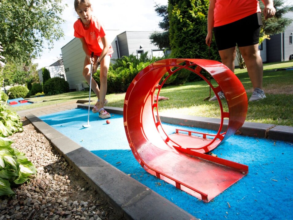Dwie osoby grają w mini golfa na zewnętrznym polu z przeszkodą w kształcie czerwonej pętli w zielonym otoczeniu.