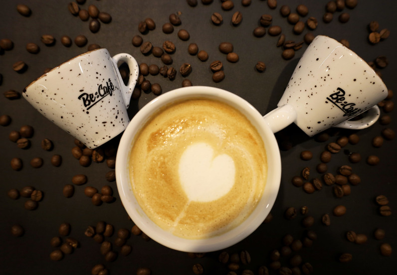 Šálka kapučína s penou v tvare srdca, okolo rozmiestnené kávové zrná a dve prázdne bodkované šálky Be:Café