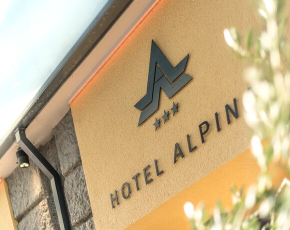 Metall-Logo mit Bergsymbol, drei Sternen und HOTEL ALPIN auf warmer Fassade, Steineingang und dezenter Vordachbeleuchtung
