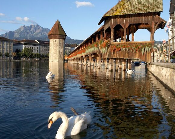Historische gedeckte Holzbrücke über dem ruhigen See mit Blumenkästen, Uferpromenade, Schwänen und Alpenpanorama