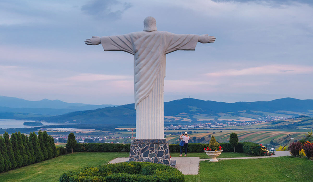 Hilltop open-armed statue overlooking a manicured garden, panoramic valley and lake at sunset; a visitor photographing the view.