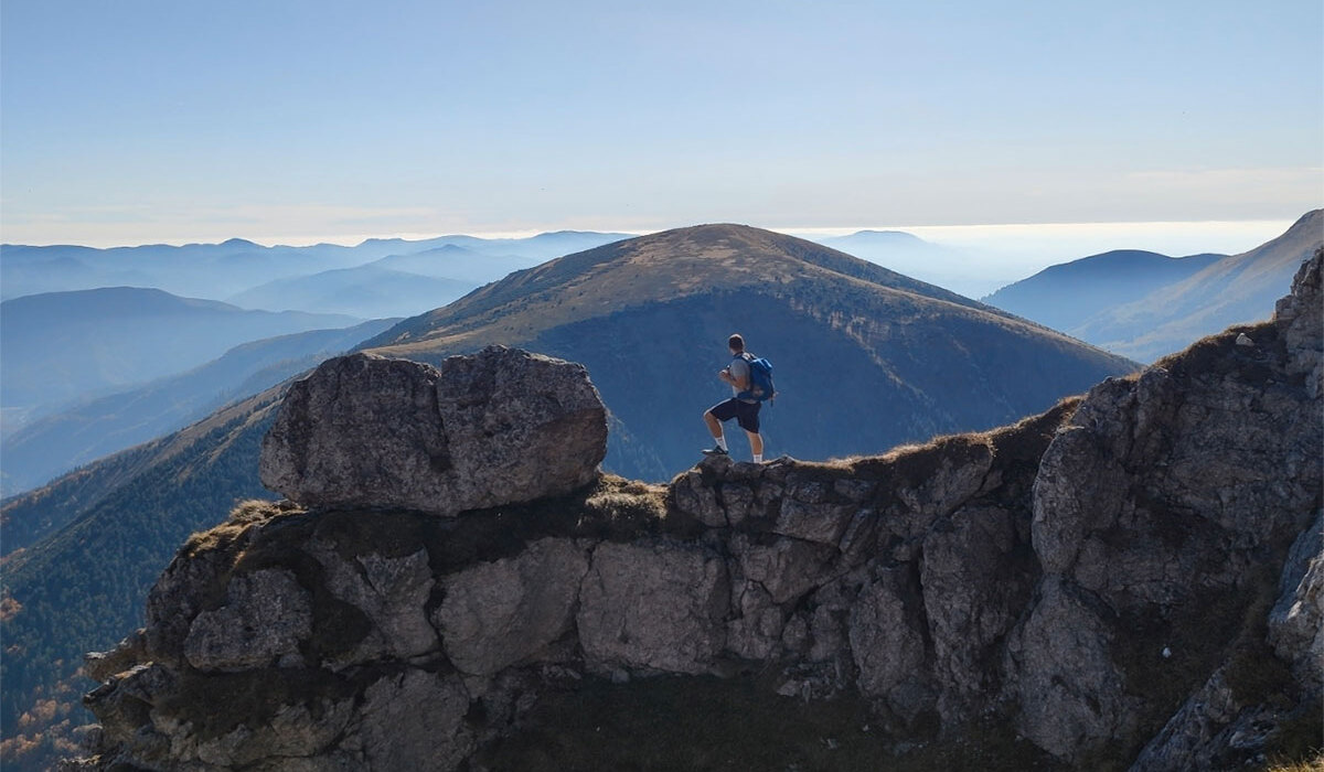 Solo hiker with backpack stands on a rocky ridge, overlooking layered blue mountain peaks under a clear sky.