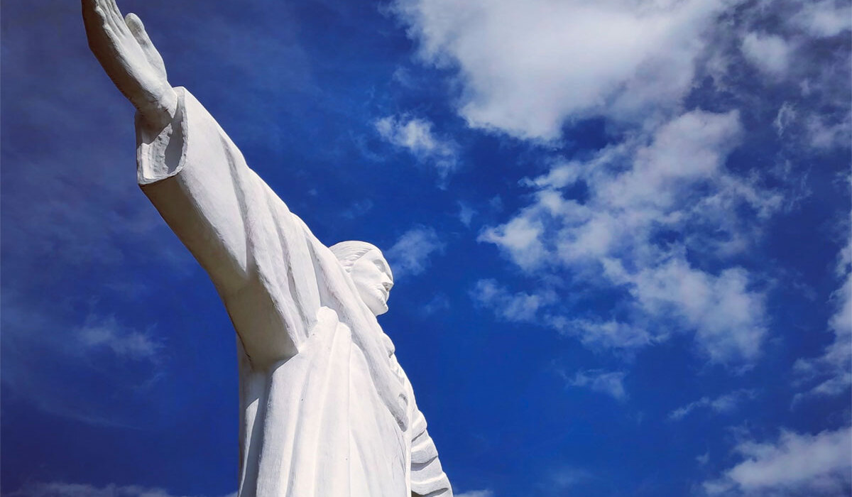Towering white Christ statue with outstretched arms framed against a bright blue sky with clouds