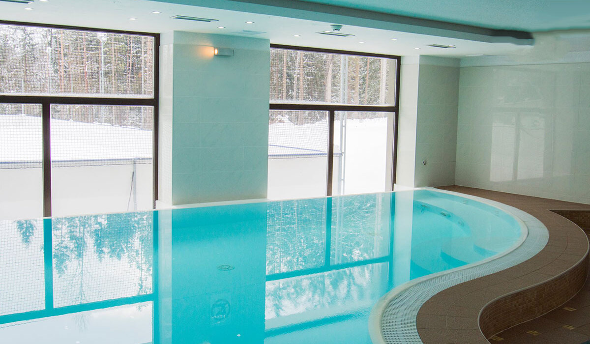 Indoor heated pool with curved tiled deck, turquoise water and large windows framing a snowy forest view.