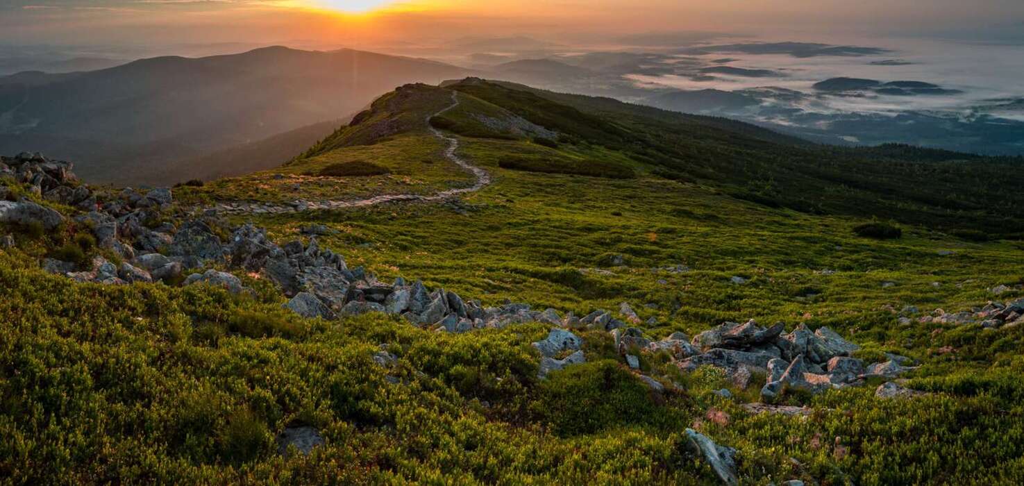 Východ slnka nad horským hrebeňom s kľukatou turistickou cestou, skalnatým predpolím a údoliami zahalenými hmlou, pokojná horská panoráma