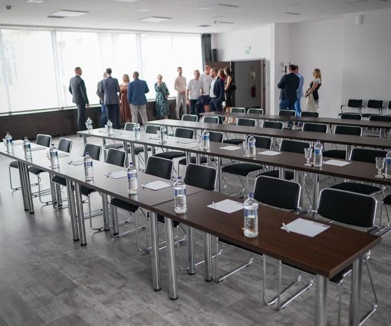 Svetlá hotelová konferenčná miestnosť s radmi stolov, fľaškovou vodou, papierom na písanie a hosťami pri okne