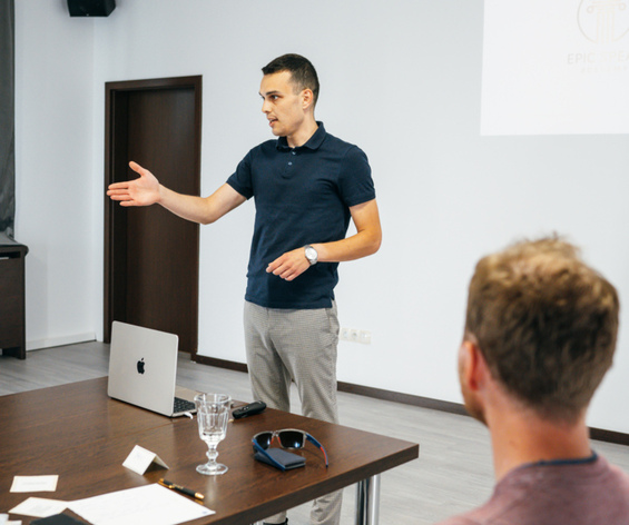Konferenčná miestnosť hotela: prednášajúci pri projekčnom plátne, stôl s notebookom, pohár vody a účastníci