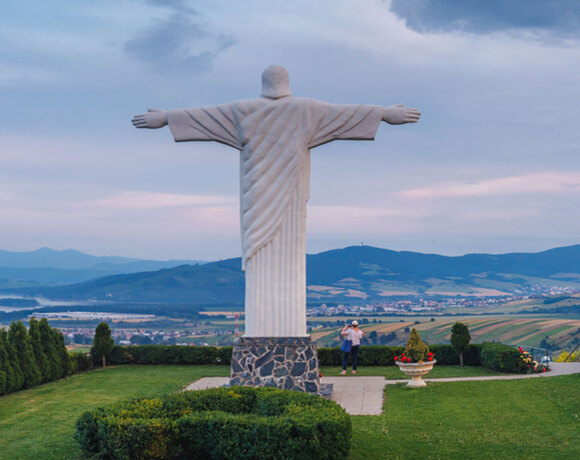 Hilltop open-armed statue overlooking a manicured garden, panoramic valley and lake at sunset; a visitor photographing the view.