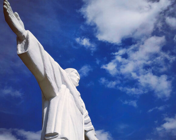 Towering white Christ statue with outstretched arms framed against a bright blue sky with clouds