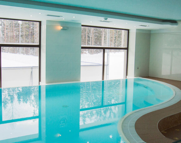 Indoor heated pool with curved tiled deck, turquoise water and large windows framing a snowy forest view.