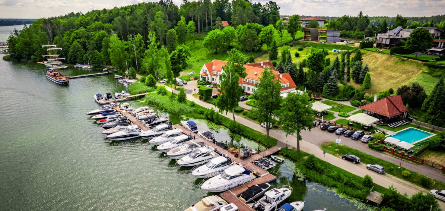 Marina z jachtami i zielonymi terenami w tle, z budynkiem hotelowym i basenem zewnętrznym.