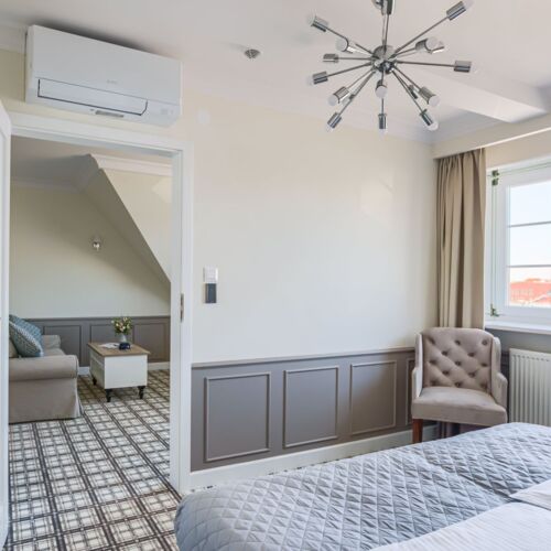 Sypialnia apartamentu z dużym łóżkiem, fotelem i widokiem na salon z kanapą i stołem