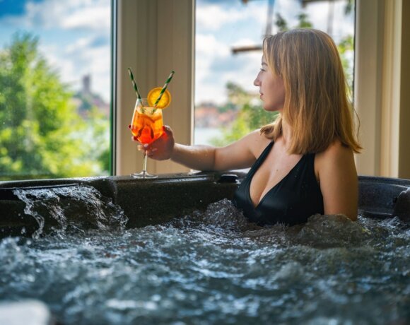 Kobieta w stroju kąpielowym z drinkiem w ręku, siedząca w jacuzzi z oknami wychodzącymi na jezioro