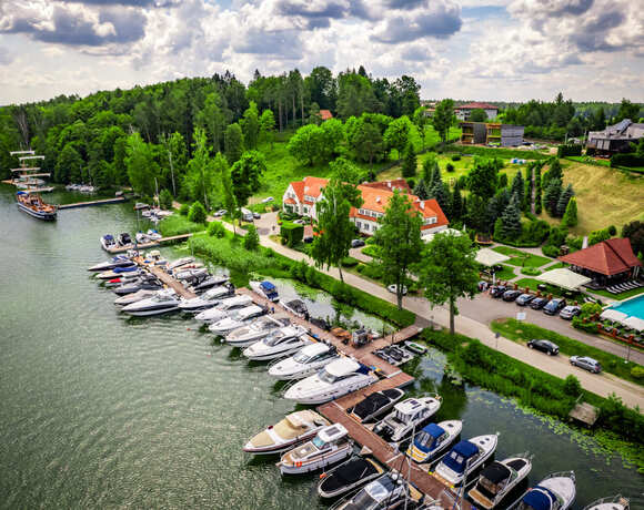 Marina z jachtami i zielonymi terenami w tle, z budynkiem hotelowym i basenem zewnętrznym.