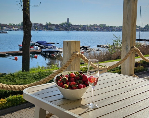 Kieliszek prosecco z truskawką obok miski pełnej truskawek na tarasie z widokiem na jezioro i miasto