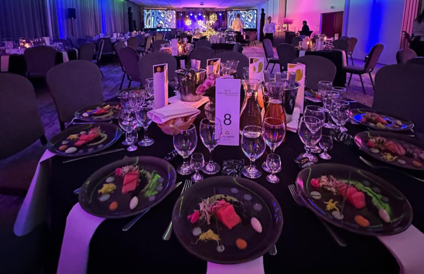 Banketttisch gedeckt für Gala-Dinner mit angerichteten Vorspeisen, mehreren Weingläsern, Blumen und violetter Bühnenbeleuchtung.