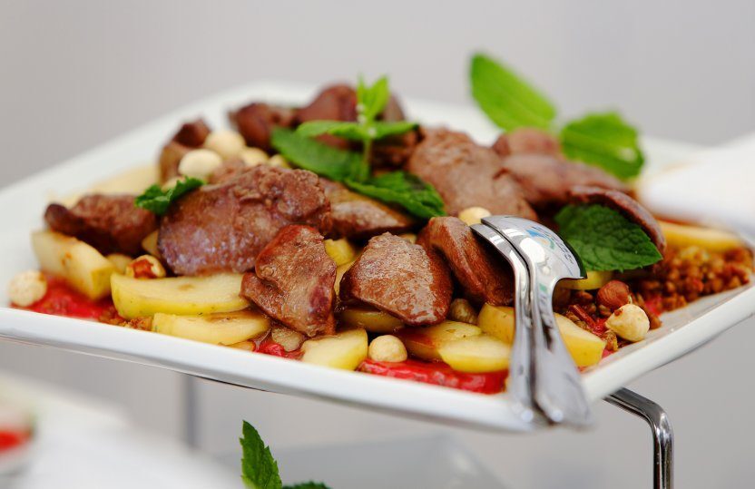 Gebratene Fleischstücke auf sautierten Kartoffelscheiben mit Haselnüssen, Minze und Servierzange auf weißer Buffetplatte.