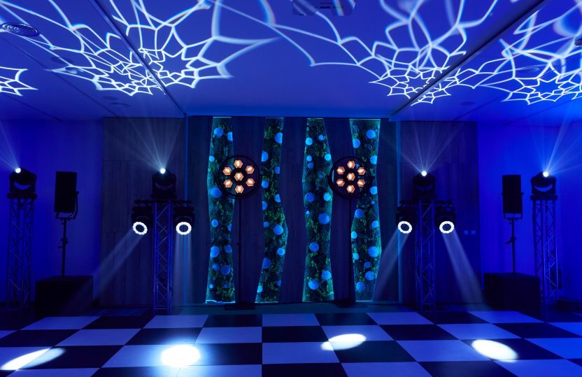 Veranstaltungsraum mit schwarz-weißem Tanzboden, blauem Ambientlicht, dekorativen Wandpaneelen und Scheinwerfern, die Muster an die Decke projizieren