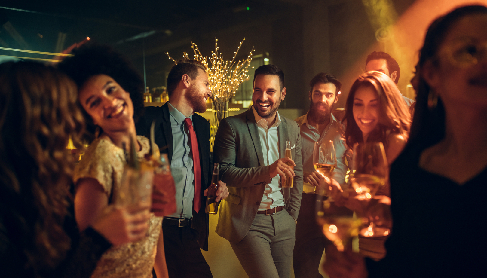 Lebhafte Hotel-Cocktailparty in gedämpfter Lounge — Gäste in Abendgarderobe lachen und halten Wein, Cocktails und Bier an der stimmungsvoll beleuchteten Bar.