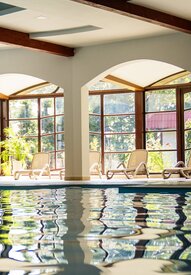 Kryty basen hotelowy z łukowymi przeszklonymi oknami, nasłonecznionymi leżakami, roślinami i drewnianymi belkami sufitowymi