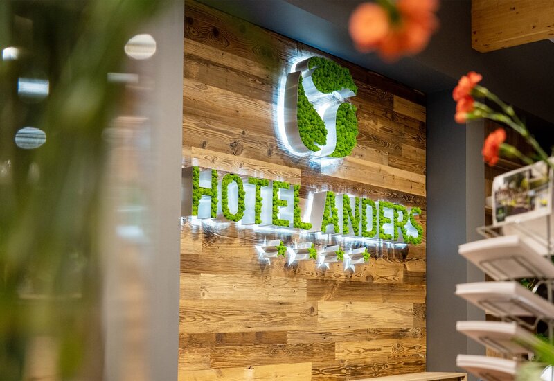 Beleuchtetes 'HOTEL LANDERS'-Logo mit grünem Moos und vier Sternen an warmer Holz-Rezeption, Prospektständer und Blumen im Vordergrund.