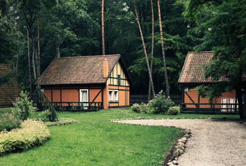 Fachwerk-Ferienhäuschen mit Ziegeldächern im Kiefernwald, mit Rasenfläche und Kiesweg.