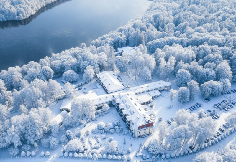 Seehotel mit kreuzförmig angeordnetem Hauptgebäude, schneebedecktem Innenhof, umgeben von frostigem Wald und Parkplätzen am Ufer.