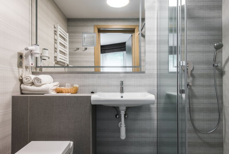 Modernes Hotelbad mit wandhängendem Waschbecken, Glasdusche, Handtuchheizkörper, gestapelten Handtüchern, Haartrockner und Kosmetikkorb