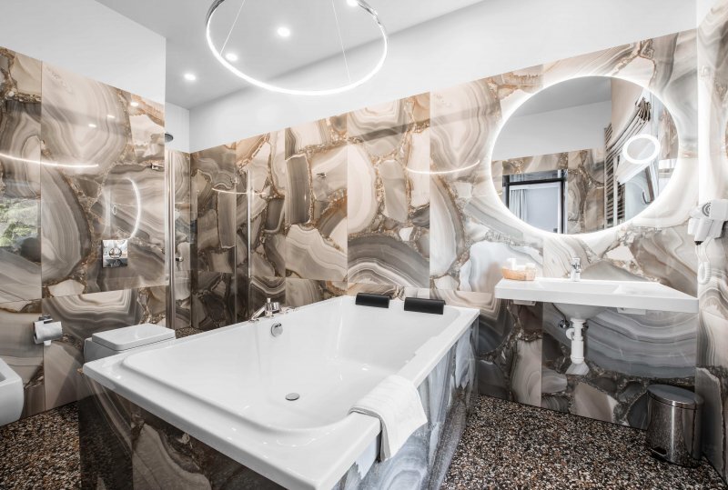 Luxury hotel bathroom with deep soaking tub, marble-patterned wall tiles, backlit round mirror, walk-in shower and modern fixtures