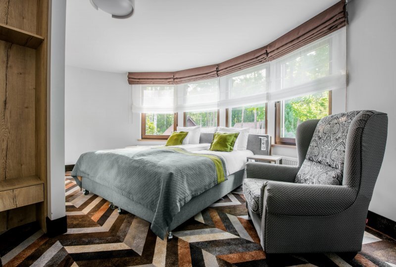 Ecksuite mit geschwungenen Panoramafenstern und Blick ins Grüne, Kingsize-Bett mit grauer Bettwäsche und grünen Kissen, gemütlicher Sessel.