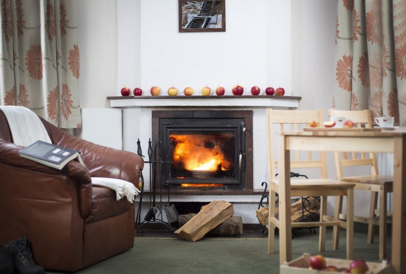 Gemütliche Lounge mit brennendem Holzofen, Ledersessel, Esstisch, Holzscheiten und Äpfeln auf dem Kaminsims.