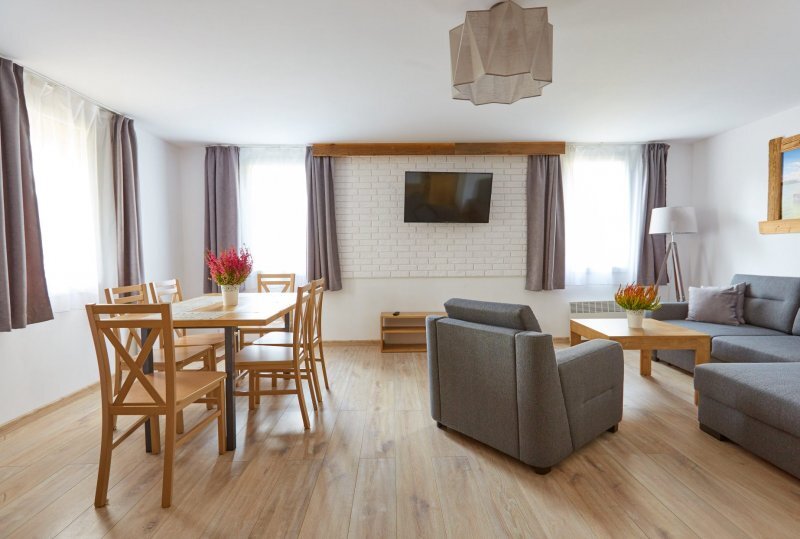 Geräumiger Wohnbereich der Suite mit Holz-Esstisch, grauem Sofa und Sessel, wandmontiertem Fernseher, viel Tageslicht und Holzfußboden