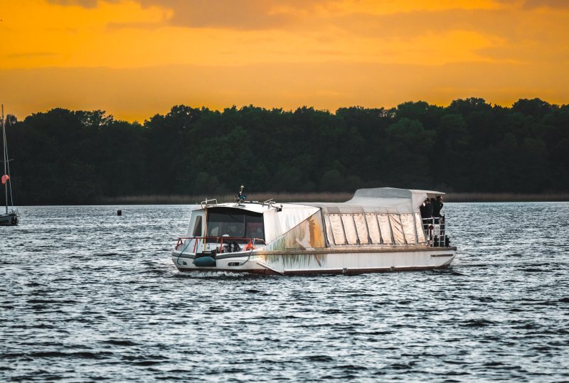 Ausflugsboot bei Sonnenuntergang mit überdachter Kabine und offenem Heckdeck, Gäste an Bord, fährt über ruhigen See vor bewaldetem Ufer