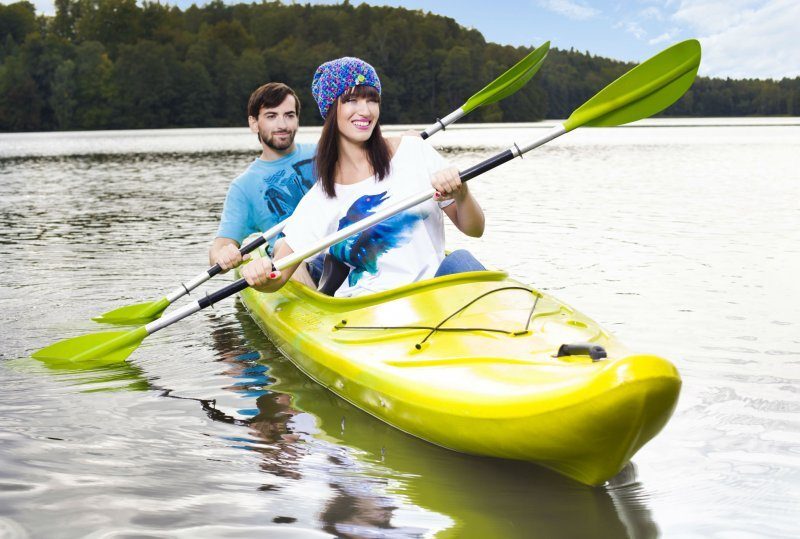 Paar paddelt im gelben Zweierkajak auf ruhigem See vor bewaldetem Ufer — Hotel-Kajakverleih und geführte Tour.