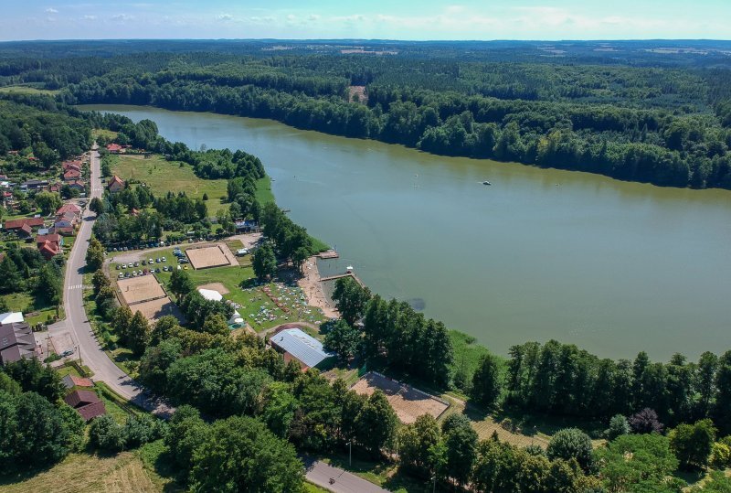 Uferbereich mit Sandstrand, Steg und grüner Liegewiese, Beachvolleyballfeldern, Dorf am See und dichtem Wald im Hintergrund
