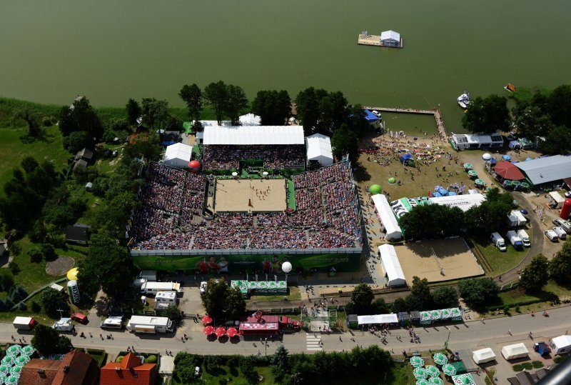 Seeseitige Beachvolleyball-Arena mit vollen Tribünen, Sandplatz, Festivalständen, Badebereich und Steg