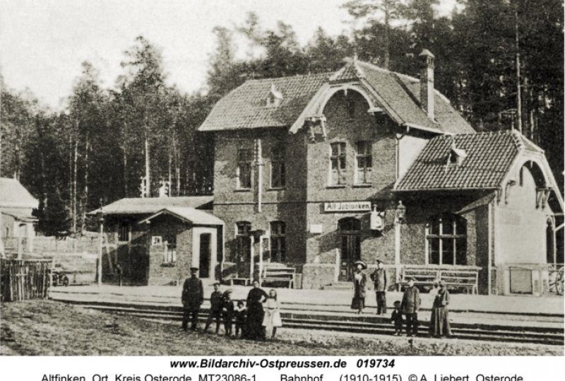 Historischer Ziegelbahnhof mit Ziegeldach und geschwungenen Giebeln, Bahnsteig mit Reisenden, Wald im Hintergrund.